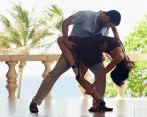 Un uomo e una donna ballano a stretto contatto in un patio assolato mentre l'uomo immerge la donna all'indietro. Le palme e il cielo azzurro incorniciano il loro momento, catturando l'energia vibrante di un corso di salsa cubana online di Freedance Academy.
