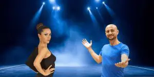 Una donna in abito nero e un uomo in camicia blu posano fiduciosi sotto le luci luminose del palcoscenico con uno sfondo blu fumoso, rappresentando Freedance Academy e le sue offerte di bachata online.